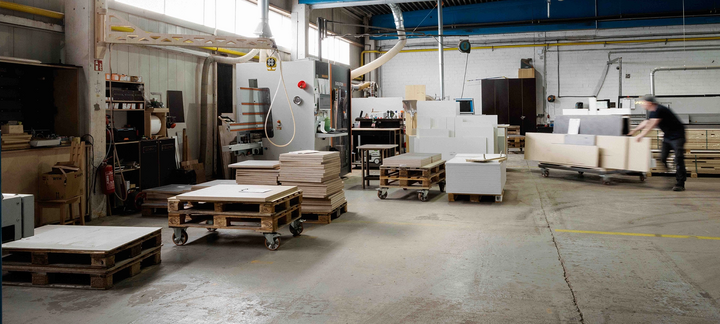 Werkhalle mit Holzpaletten, verpackten Warenstapeln und einer Person an einer Maschine.