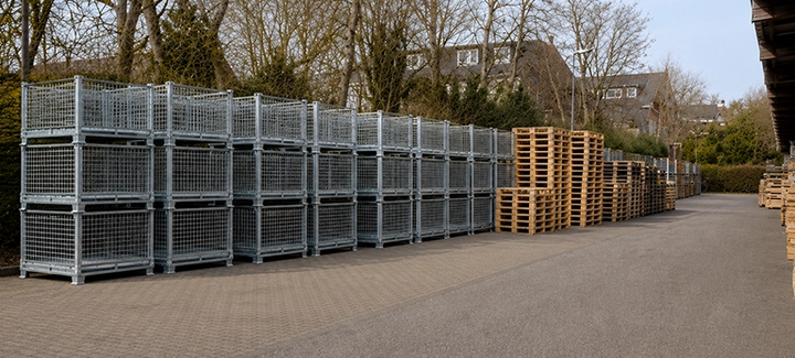 Stapelbare Metall-Gitterboxen im Außenlager neben gestapelten Holzpaletten.