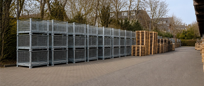 Stapelbare Metall-Gitterboxen im Außenlager neben gestapelten Holzpaletten.