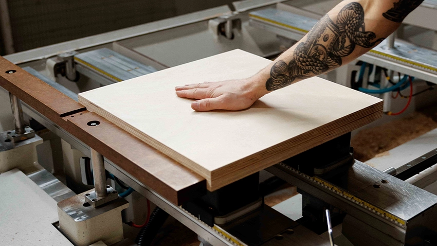Hand legt eine Holzplatte auf den Tisch einer CNC‑Fräse.