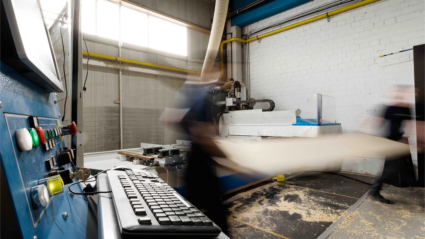 Zwei Personen bewegen eine große Holzplatte vor einer CNC‑Maschine, im Vordergrund die Tastatur der Steuerung.